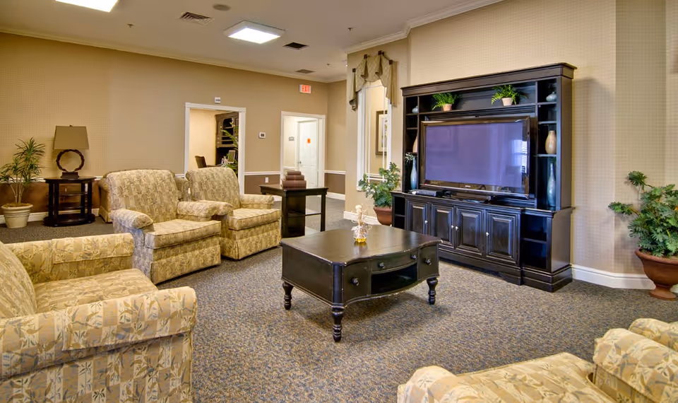 A cozy senior living facility common area with patterned armchairs arranged around a dark wooden coffee table. A large dark wooden entertainment center with a flat-screen TV and decorative plants is against the wall. The room has beige walls, carpeted flooring, and several potted plants. There are doorways leading to other rooms in the background.