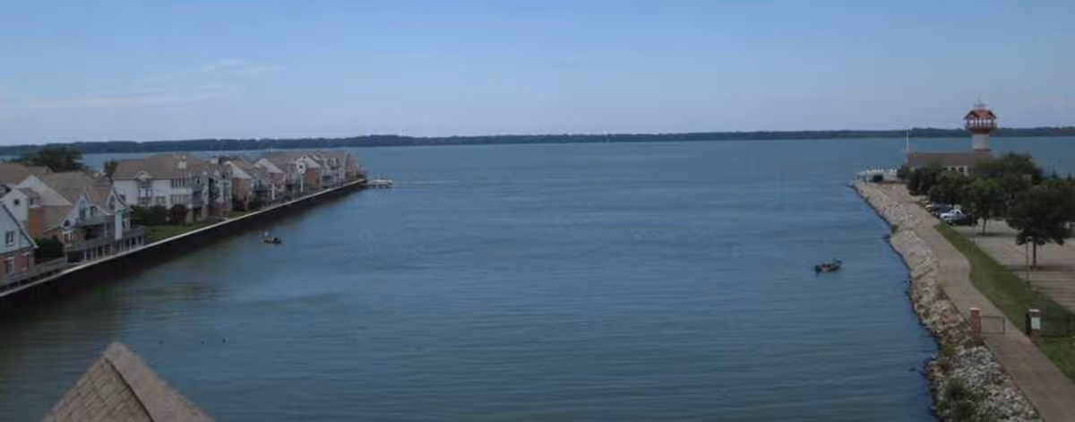Wide view of a calm bay with row houses along the left shoreline, a lighthouse and rocky breakwater on the right, and small boats on the water.