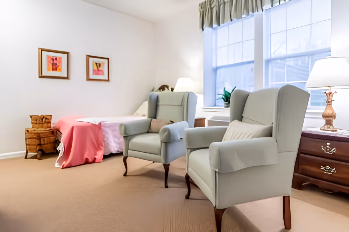 A cozy bedroom with two light gray armchairs in the foreground, a bed with a pink blanket against the wall, two framed pictures above the bed, a wooden nightstand with a lamp, and large windows with green curtains letting in natural light.