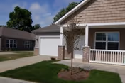 Exterior view of a single-story residential building with a garage, a small tree planted in a mulched area, and a porch with white railing. Another similar building is visible in the background under a partly cloudy sky.