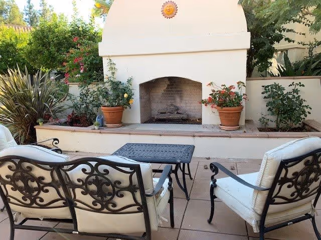 Outdoor patio area with cushioned wrought iron chairs and a matching table facing a built-in outdoor fireplace surrounded by potted plants and greenery.