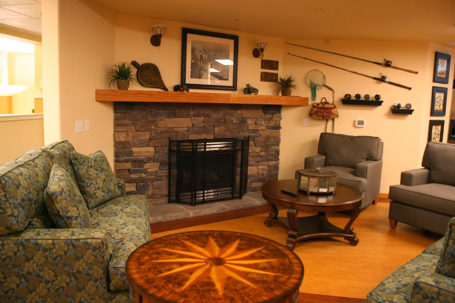 Cozy living room with a stone fireplace, wooden mantel decorated with plants, a framed photo, and fishing gear mounted on the wall. The room has patterned green sofas, two gray armchairs, and two wooden coffee tables with decorative items on them.