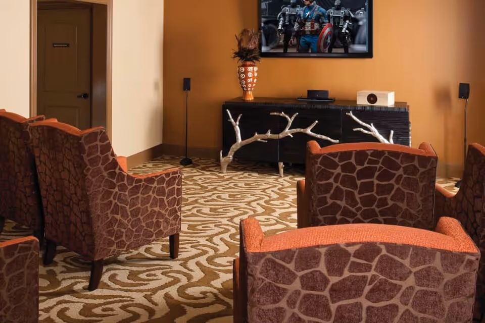 A cozy living room area with several patterned armchairs arranged facing a wall-mounted TV displaying a superhero scene. The room has a patterned carpet, a black cabinet with decorative antler handles, and a vase with feathers on top. The walls are painted in warm tones, and a door labeled 'Restroom' is visible in the background.
