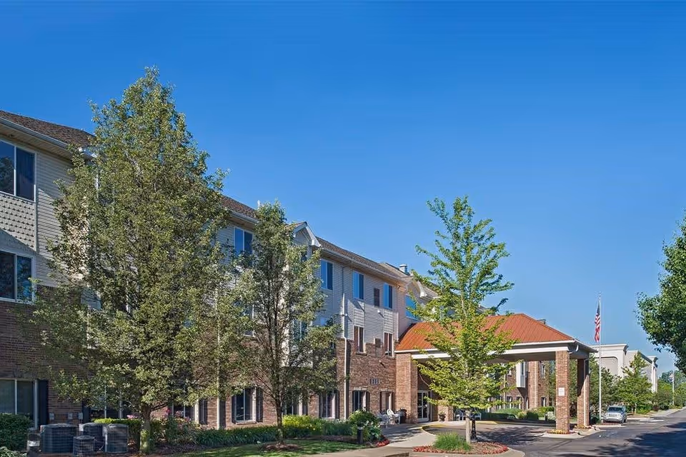Exterior view of a multi-story senior living facility with brick and siding facade, surrounded by trees and landscaping under a clear blue sky. There is a covered entrance with a red roof and an American flag visible in the background.