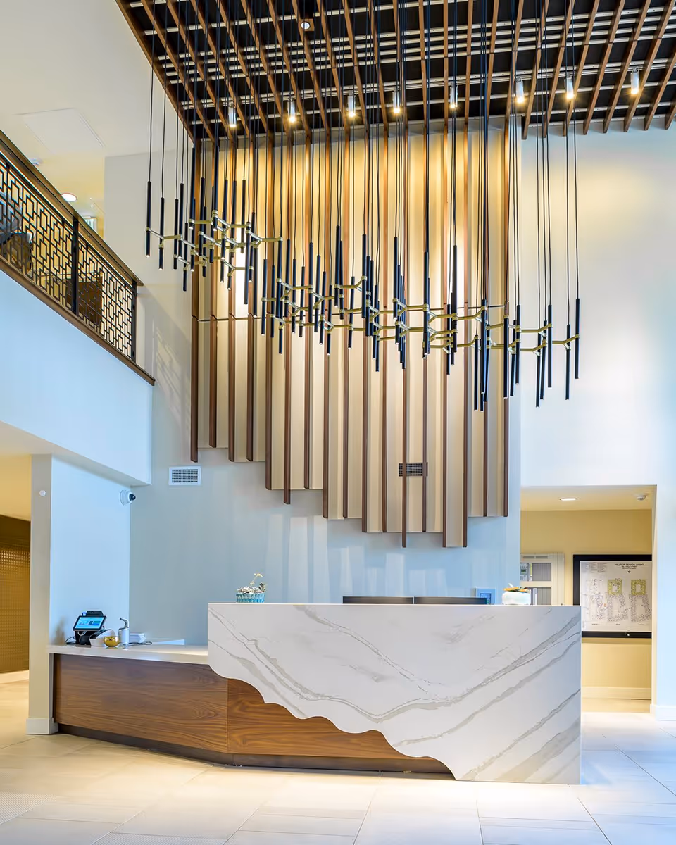 Modern reception desk area in a senior living facility with a white marble front and wooden base. Above the desk, there is a large decorative wooden panel and a contemporary chandelier with multiple hanging black and gold light fixtures. The space has high ceilings and light-colored tiled floors.