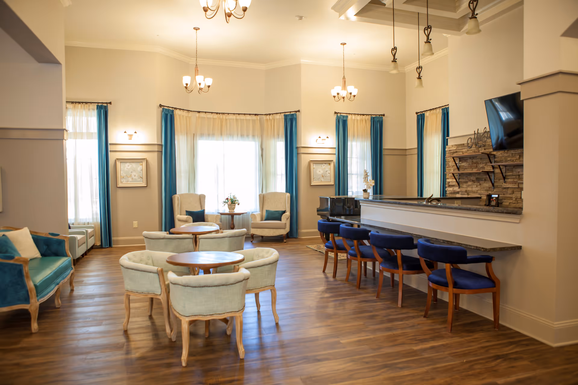 Bright, well-appointed senior living common lounge with round tables, upholstered chairs, a bar counter with stools, and large windows with blue curtains.