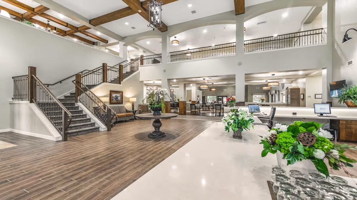 Spacious and well-lit interior of a senior living facility featuring a large open lobby area with wooden flooring, a central round table with a floral arrangement, a staircase with wooden handrails leading to an upper floor balcony, multiple seating areas with chairs and tables, and a reception desk with computer monitors and additional floral decorations.