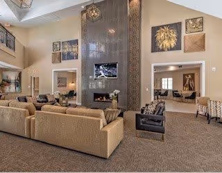 Spacious living room area with beige sofas, armchairs, and a large decorative fireplace with a mounted TV above it. The room features high ceilings, wall art, and an open layout connecting to another seating area in the background.
