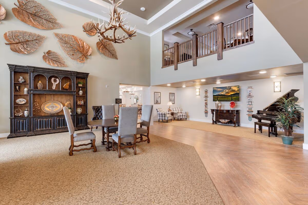 Spacious senior living facility common area with high ceilings and a second-floor balcony. The room features a round wooden table with four upholstered chairs, a large dark wood cabinet with decorative items, and a chandelier made of antlers. On the right side, there is a grand piano next to a potted plant, a console table with vases and a red decorative plate, two patterned armchairs with a lamp between them, and a wall-mounted TV displaying a scenic landscape.