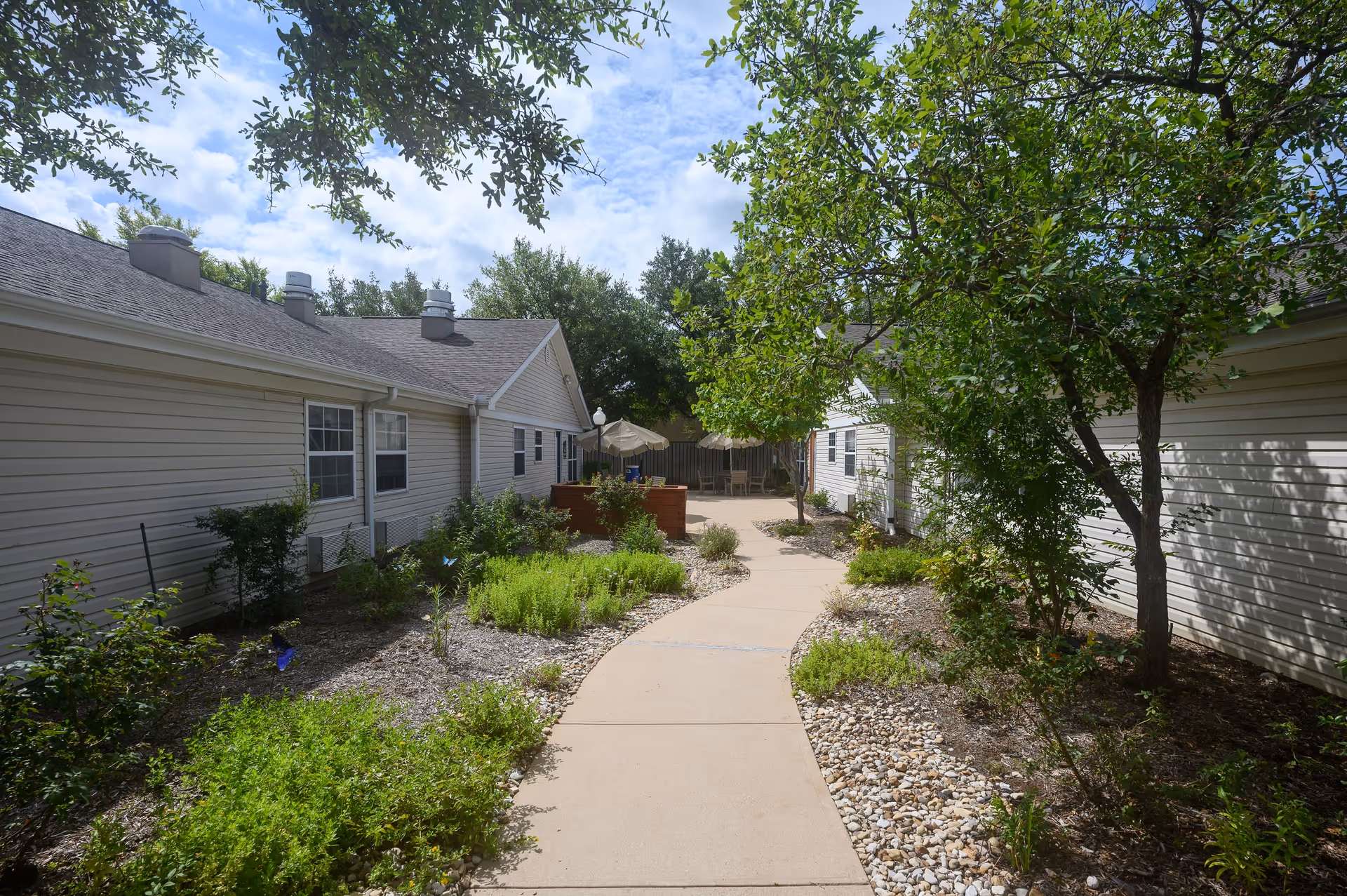 A paved walkway runs between two single-story buildings with beige siding, surrounded by landscaped garden beds with green shrubs, small plants, and trees under a partly cloudy sky.