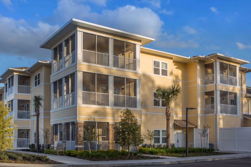 Exterior view of a three-story senior living facility building with yellow siding, multiple screened balconies, palm trees, and landscaped greenery under a partly cloudy sky.