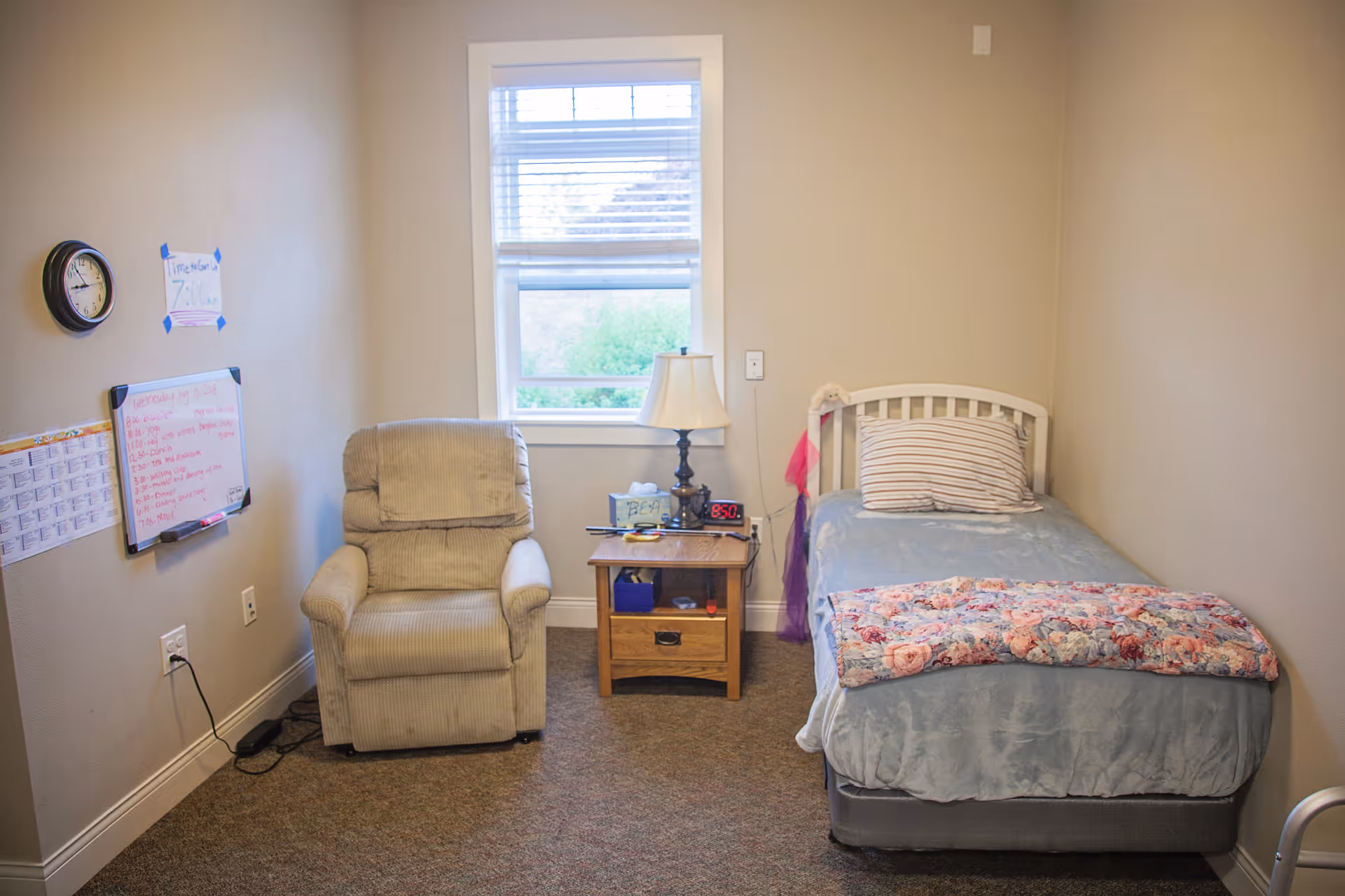 Small furnished bedroom with a single bed, recliner chair, nightstand and window in a memory care facility.