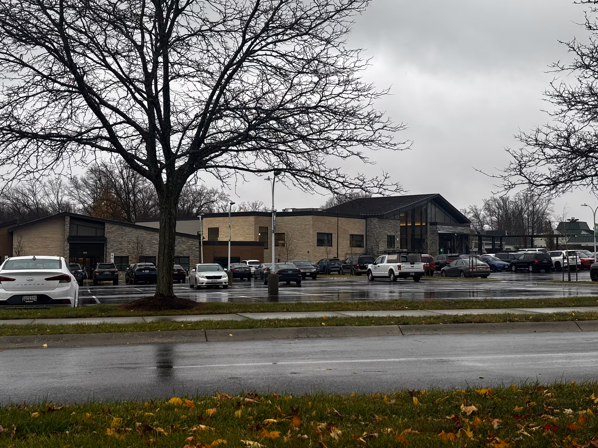 Exterior front view of a single-story senior living facility with a parking lot, cars, and leafless trees on a rainy day.