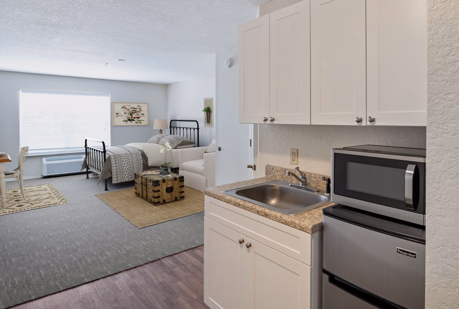 A bright and clean studio apartment featuring a small kitchenette with a sink, microwave, and mini refrigerator on the right. The living and sleeping area includes a black metal bed frame with bedding, a white armchair, a wooden chest used as a coffee table, a desk with a chair near a large window with blinds, and wall decorations including a framed picture and a small plant. The floor transitions from wood near the kitchenette to carpet in the living and sleeping area.