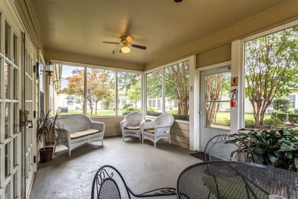 Enclosed screened porch with wicker loveseat and chairs, metal table and chairs, and large windows overlooking a landscaped courtyard.
