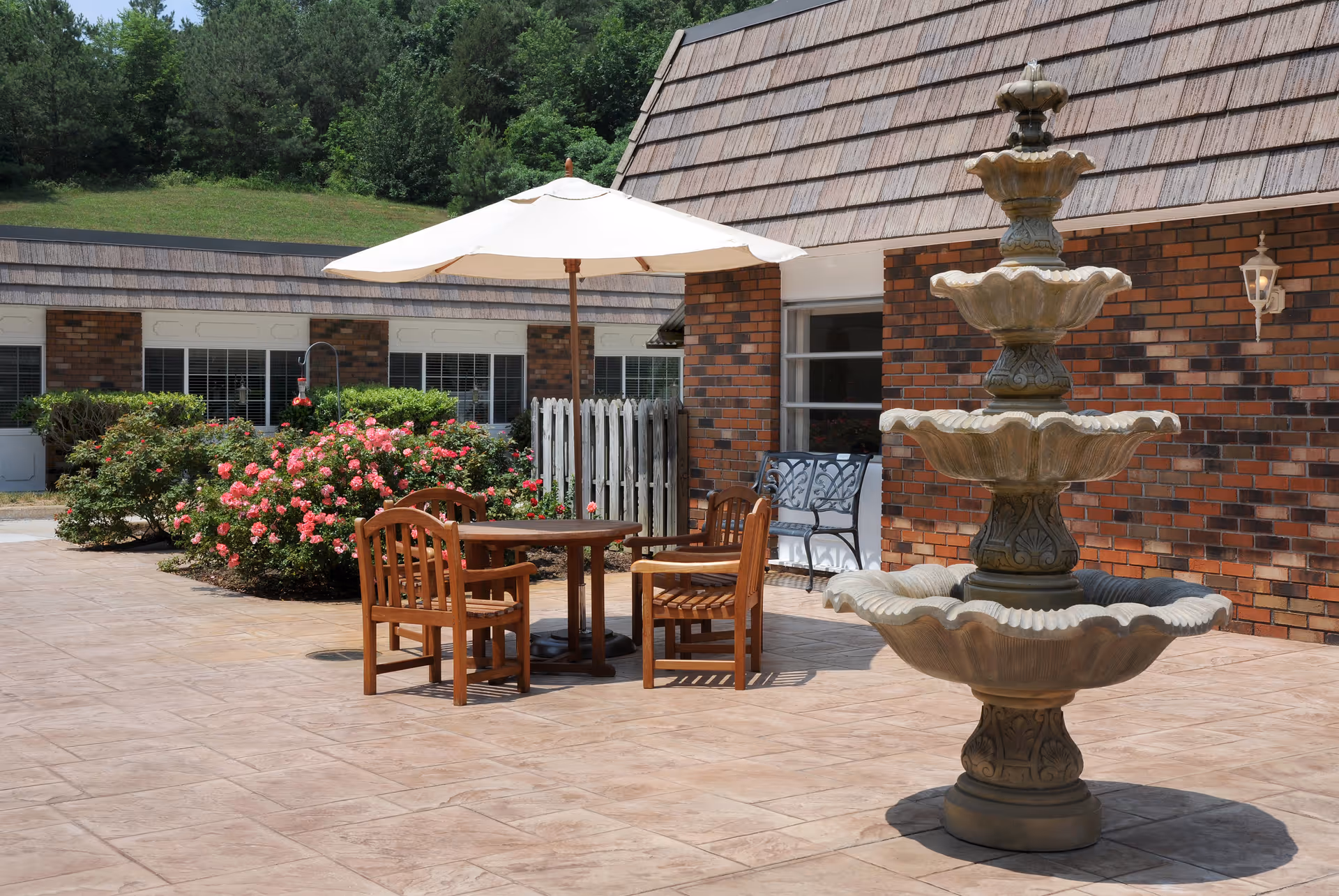 Outdoor patio area with a stone fountain in the foreground, a wooden table with four chairs under a large white umbrella, and a brick building with windows and a bench in the background. There are bushes with pink flowers and green trees behind the building.