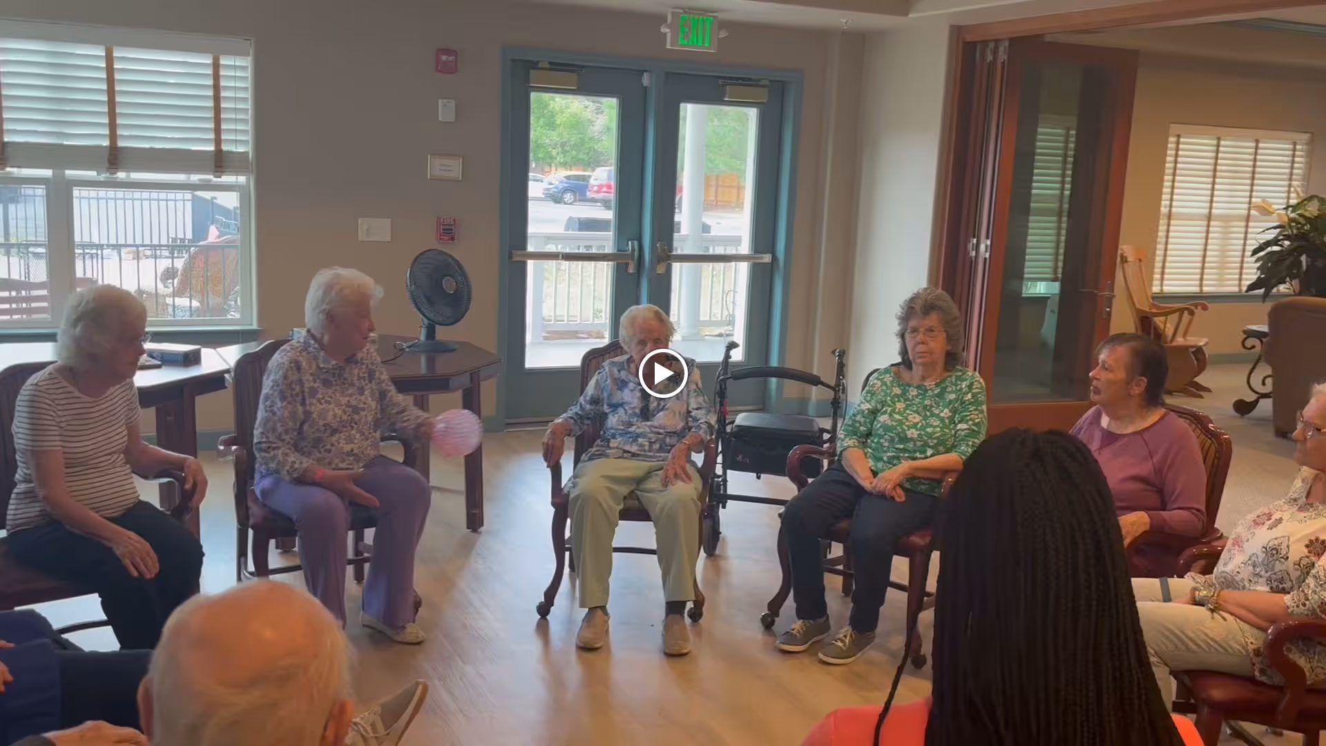 A group of elderly people sitting in a circle of chairs inside a well-lit room with large windows and double glass doors. They appear to be engaged in a group activity, with one person holding a small ball. The room has wooden flooring and some furniture, including a table with a fan on it and a walker nearby.