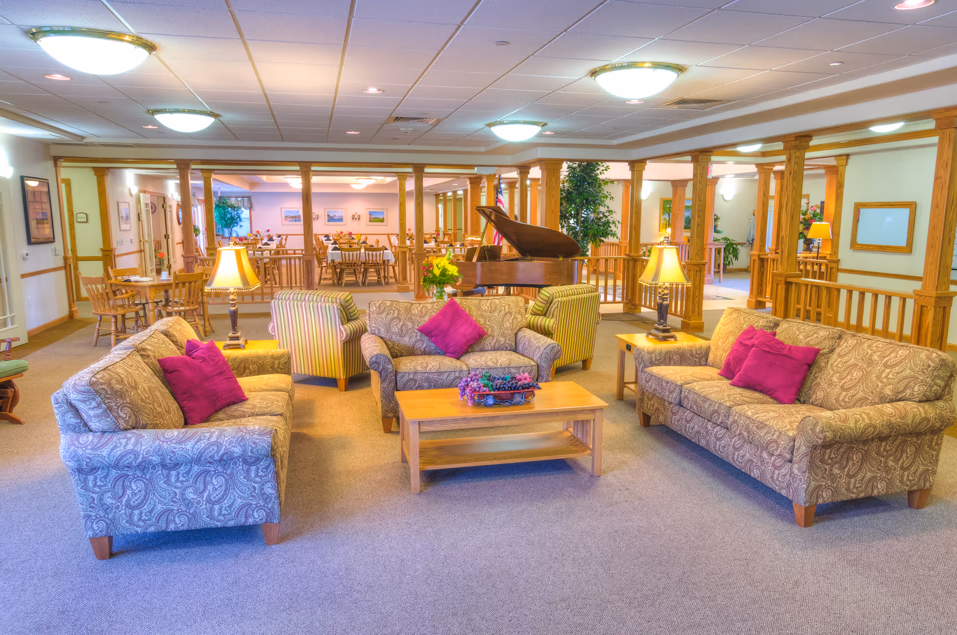 A spacious and well-lit living room area in a senior living facility with three patterned sofas adorned with purple cushions arranged around a wooden coffee table with a flower arrangement. Two striped armchairs and two table lamps on side tables flank the sofas. In the background, there is a grand piano and a dining area with wooden tables and chairs. The room features wooden columns and railings, carpeted flooring, and ceiling lights.