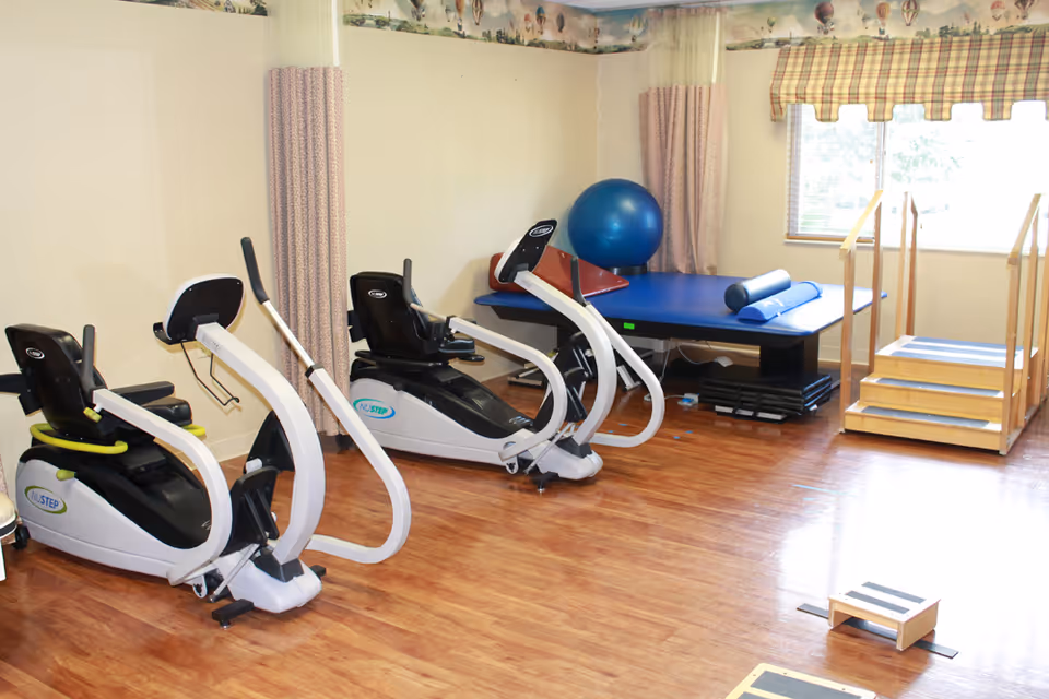 A rehabilitation exercise room with two recumbent exercise bikes, a blue exercise ball, a padded therapy table with foam rollers, and a wooden staircase with handrails for physical therapy. The room has wood flooring, light-colored walls, a window with plaid valance, and privacy curtains.