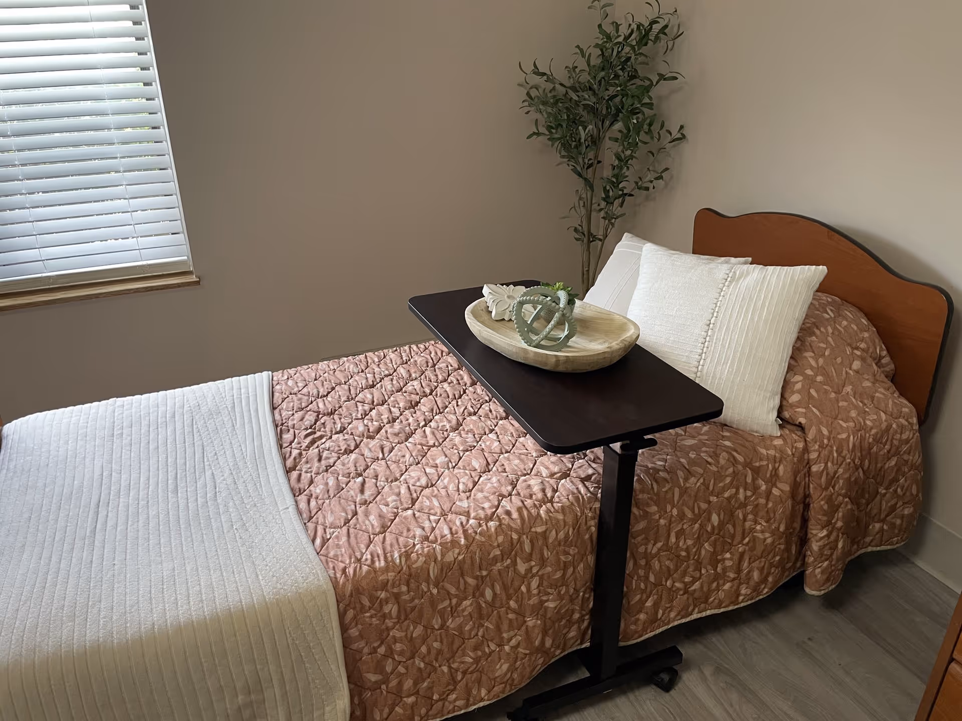 Single bed in a small room with a pink quilted bedspread, white pillows, an overbed table holding a decorative tray, a potted plant, and a window with blinds.