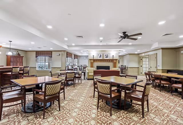 A spacious senior living facility common dining area with multiple wooden tables and chairs arranged neatly on a patterned carpet. The room features a fireplace with framed artwork above it, ceiling lights, a ceiling fan, and windows with valances allowing natural light to enter.