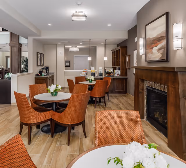 Communal dining and lounge area with round tables, orange woven chairs, a fireplace, and a beverage station in the back.