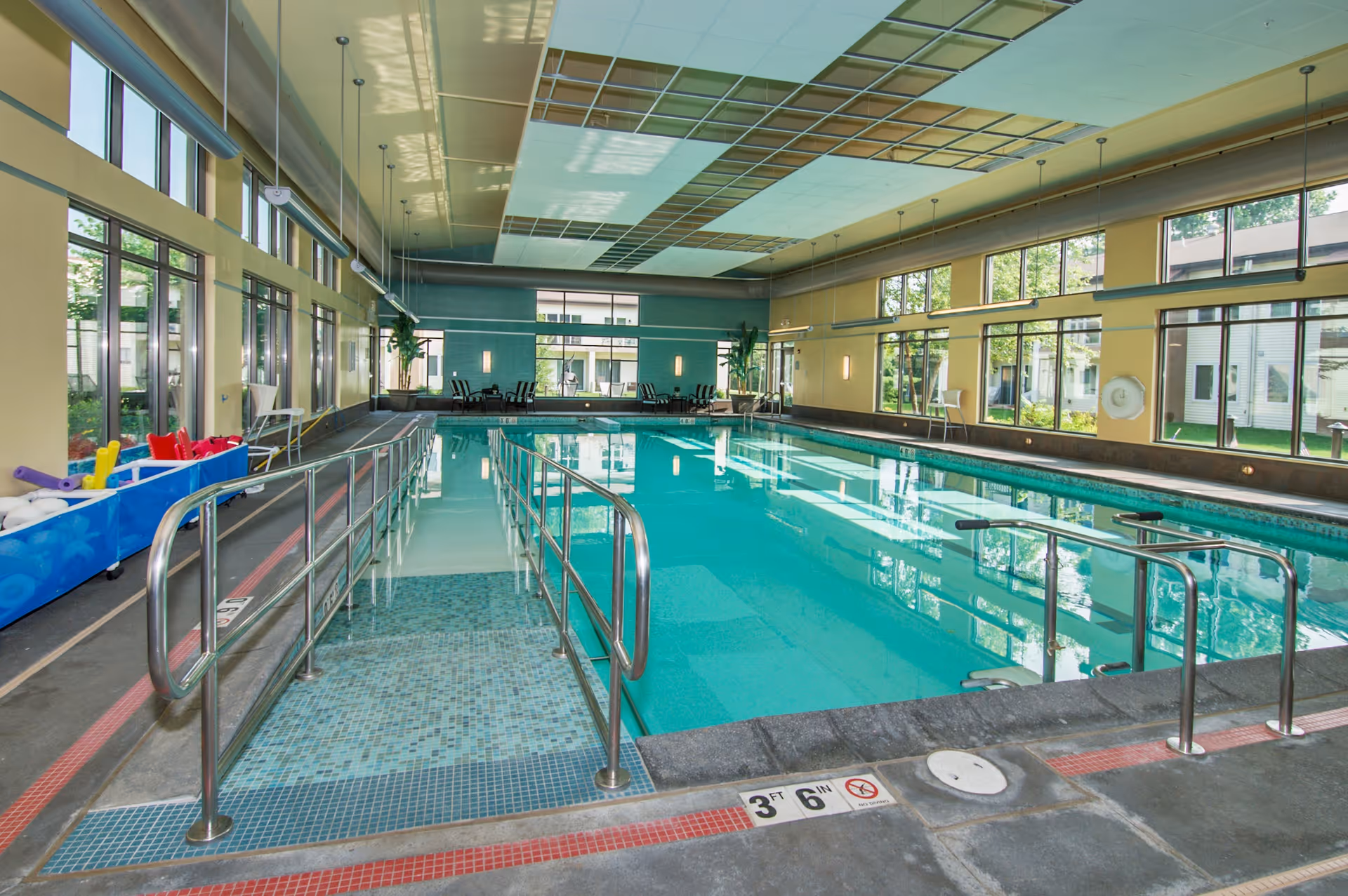 Indoor swimming pool with handrails, lounge chairs, and large windows along the sides.
