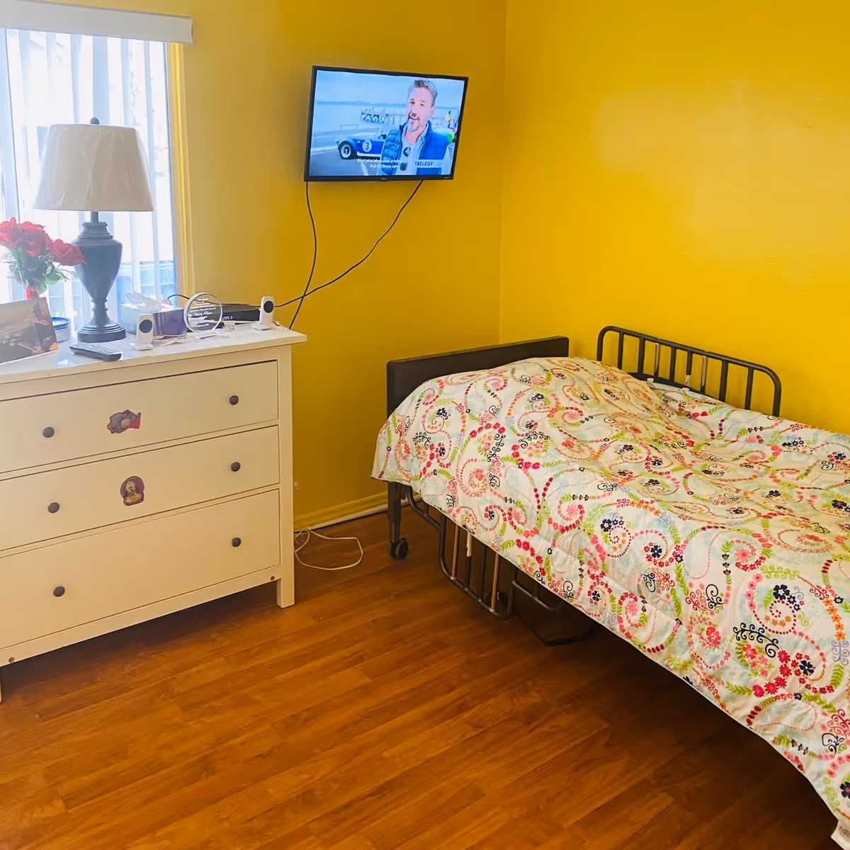 A small bedroom with yellow walls and wooden floor. There is a single bed with a colorful patterned quilt, a white dresser with a lamp, flowers, and other small items on top. A flat-screen TV is mounted on the wall above the bed, showing a man speaking.