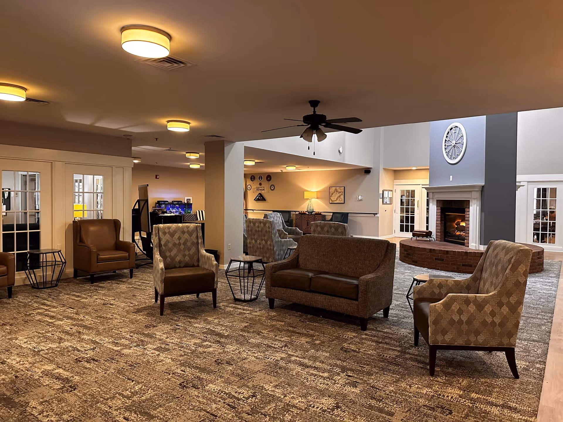 A spacious and warmly lit common area with multiple armchairs and a loveseat arranged around small tables. The room features a large brick fireplace with a clock mounted above it, a ceiling fan, and soft carpeting. In the background, there are glass-paneled doors and a fish tank, creating a cozy and inviting atmosphere.