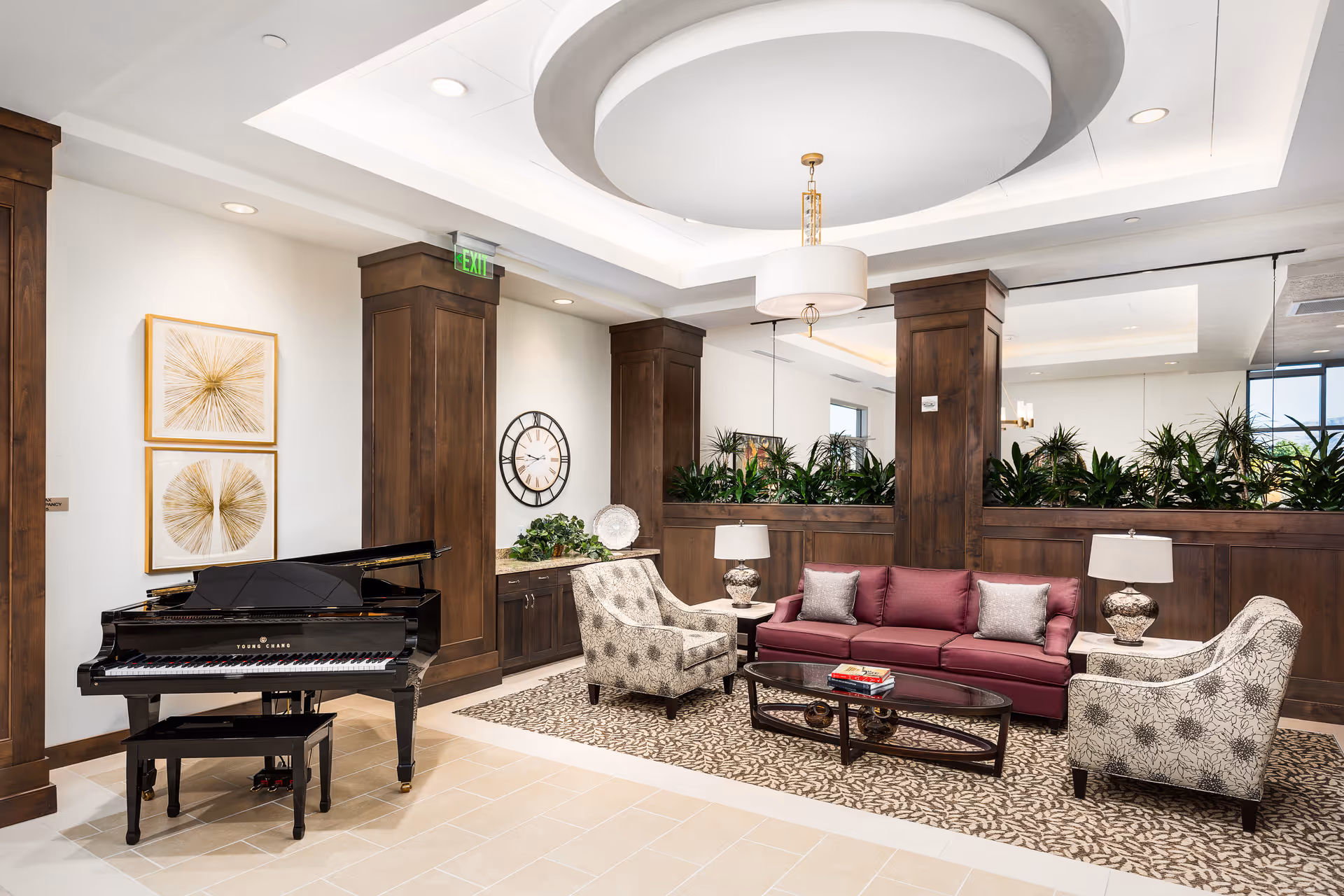 A bright and elegant living room area in Legacy Village of Sugar House featuring a black grand piano on the left, a maroon sofa with two gray pillows, two patterned armchairs, a glass coffee table with books, two table lamps, decorative plants, and wall art with a large clock and golden abstract designs.