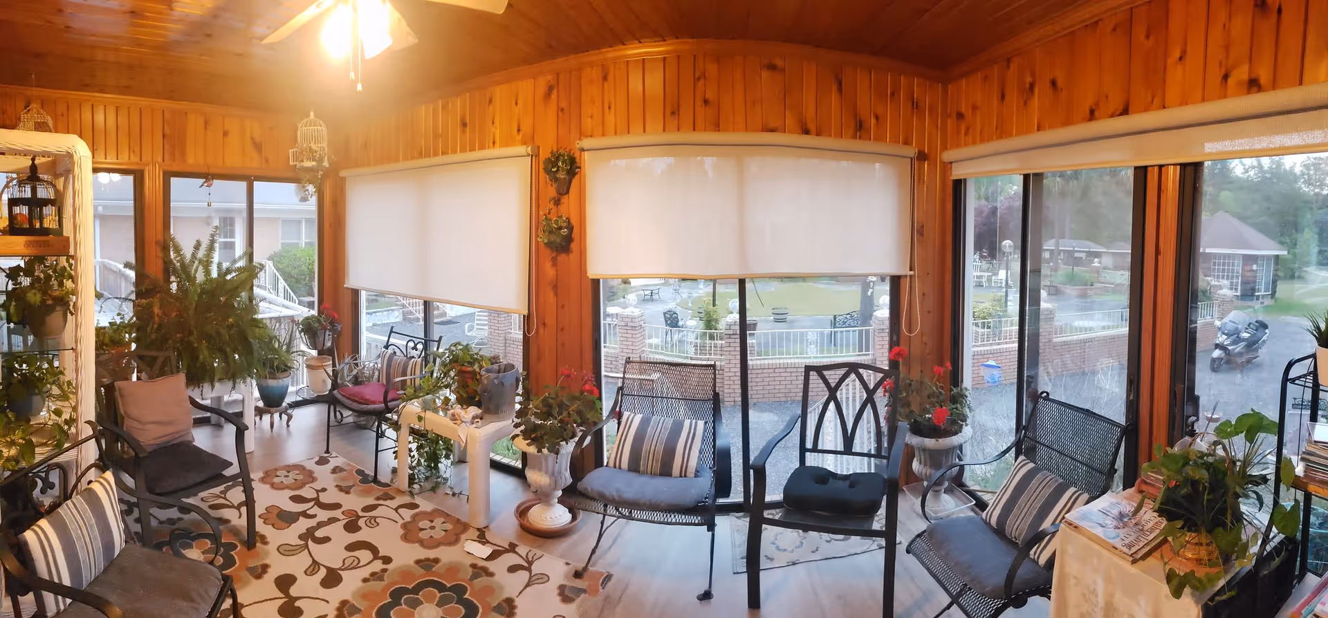 A cozy sunroom with wooden walls and ceiling, furnished with several metal chairs with cushions, a floral patterned rug, and various potted plants. Large windows with roller shades provide a view of an outdoor garden and patio area.