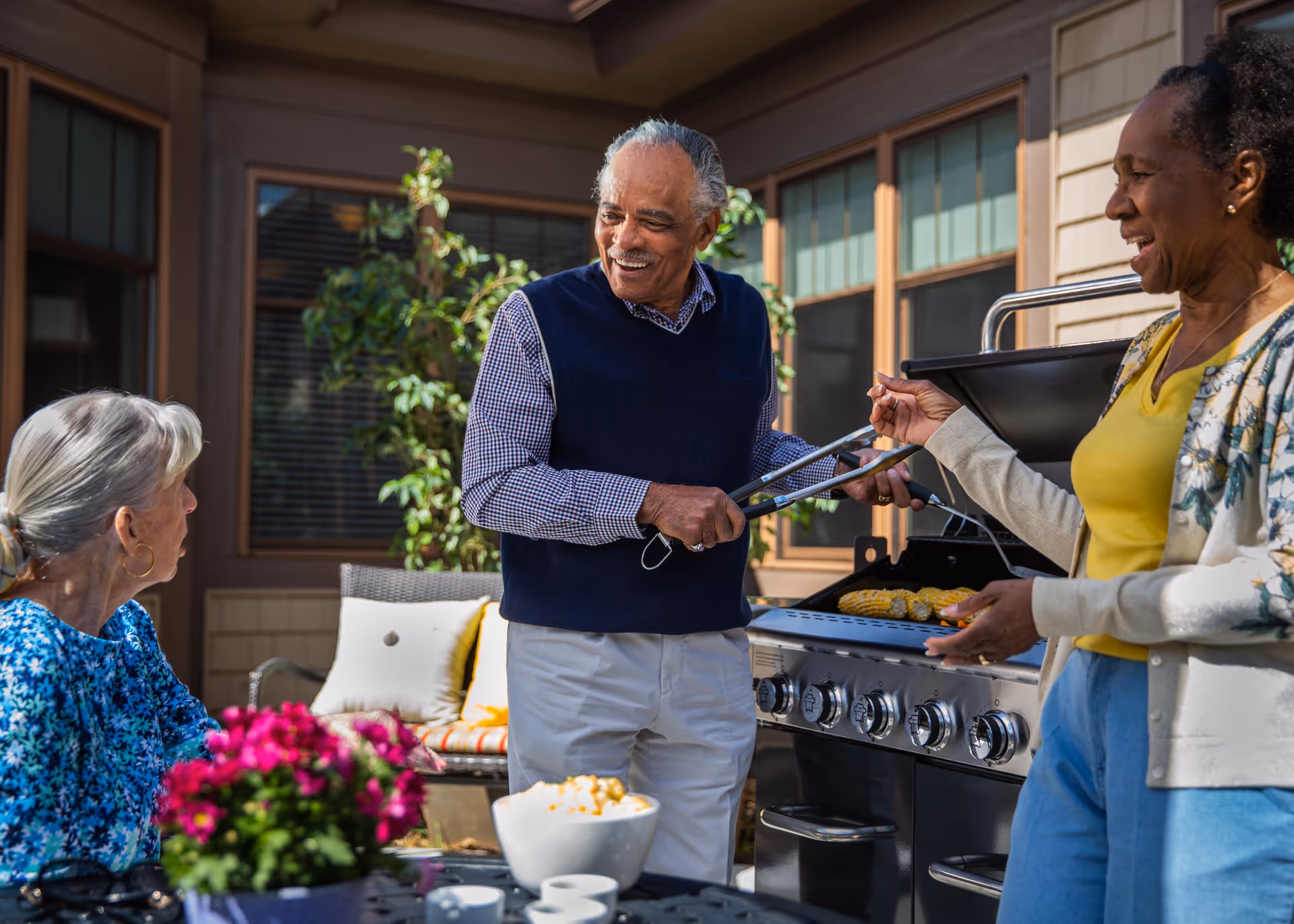 Three elderly people enjoying a barbecue outdoors on a patio. One man is holding tongs and smiling while standing near a grill with food cooking. Two women are nearby, one sitting at a table with a bowl of popcorn and flowers, and the other standing and interacting with the man. The setting is a sunny day with a building and windows in the background.