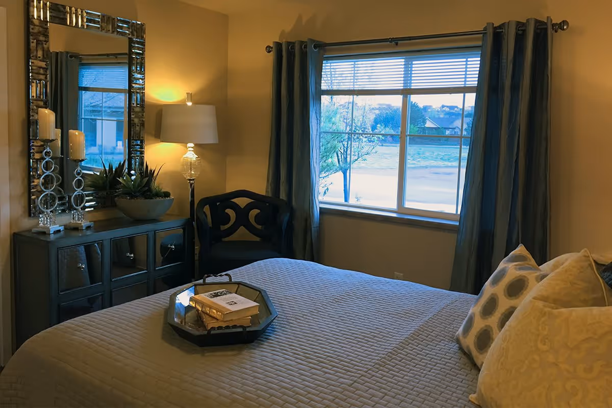 A well-decorated bedroom featuring a made bed with pillows, a dresser with mirror and lamp, a chair, and a window with curtains.