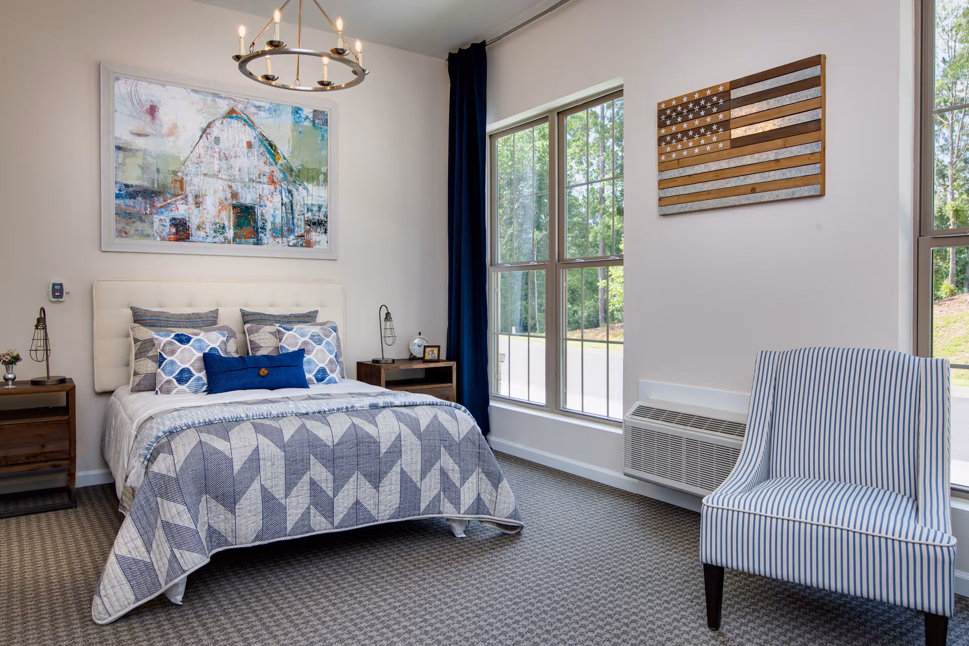 A neatly made bed with a geometric patterned quilt and multiple pillows in a bedroom. The room features two large windows with dark blue curtains, a striped armchair, two wooden nightstands with lamps, and wall art including a painting of a barn and a wooden American flag.
