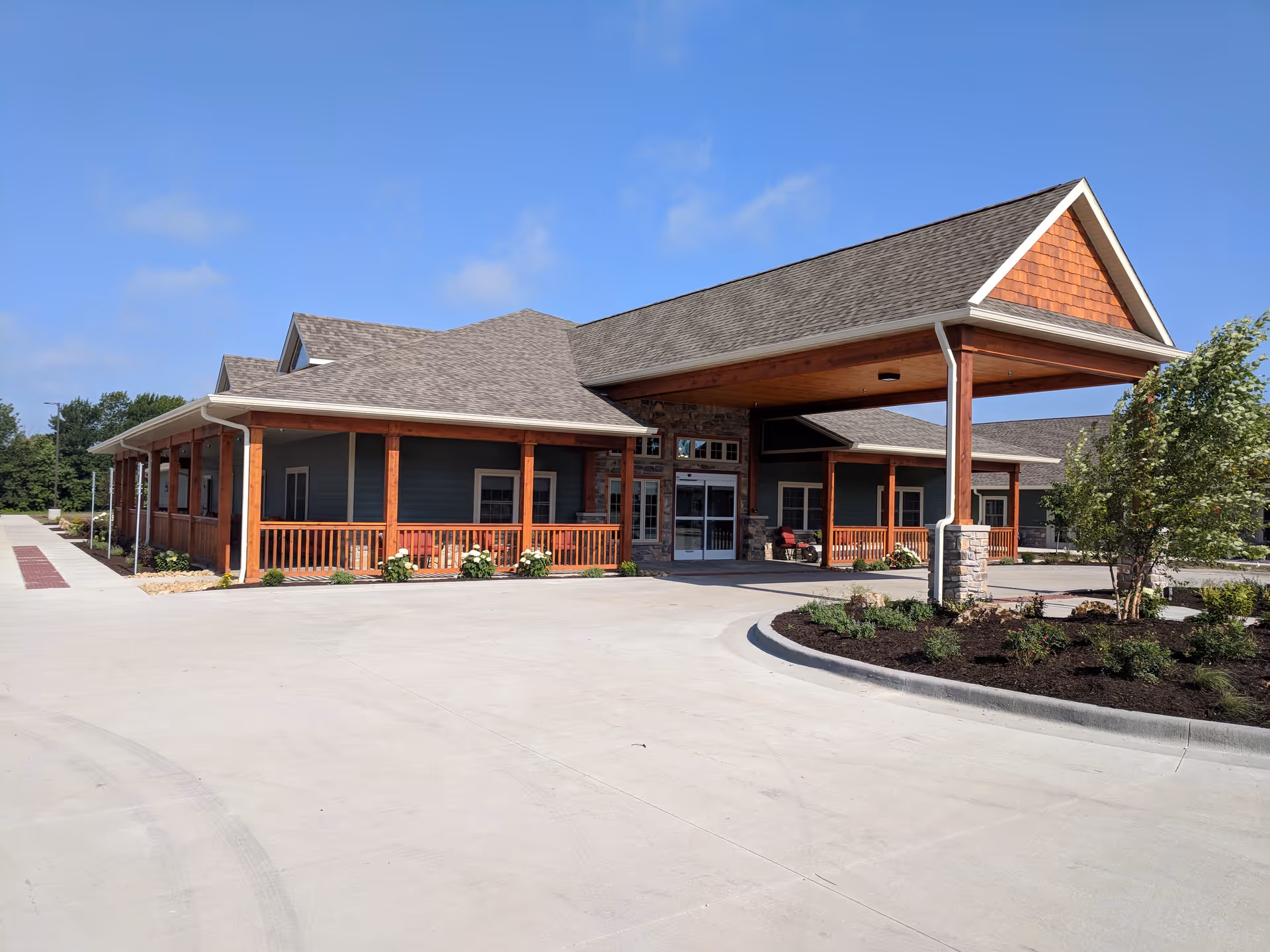 Front entrance of a single-story assisted living building with a covered porte-cochère, wooden columns, porch railings, and landscaped driveway under a blue sky.