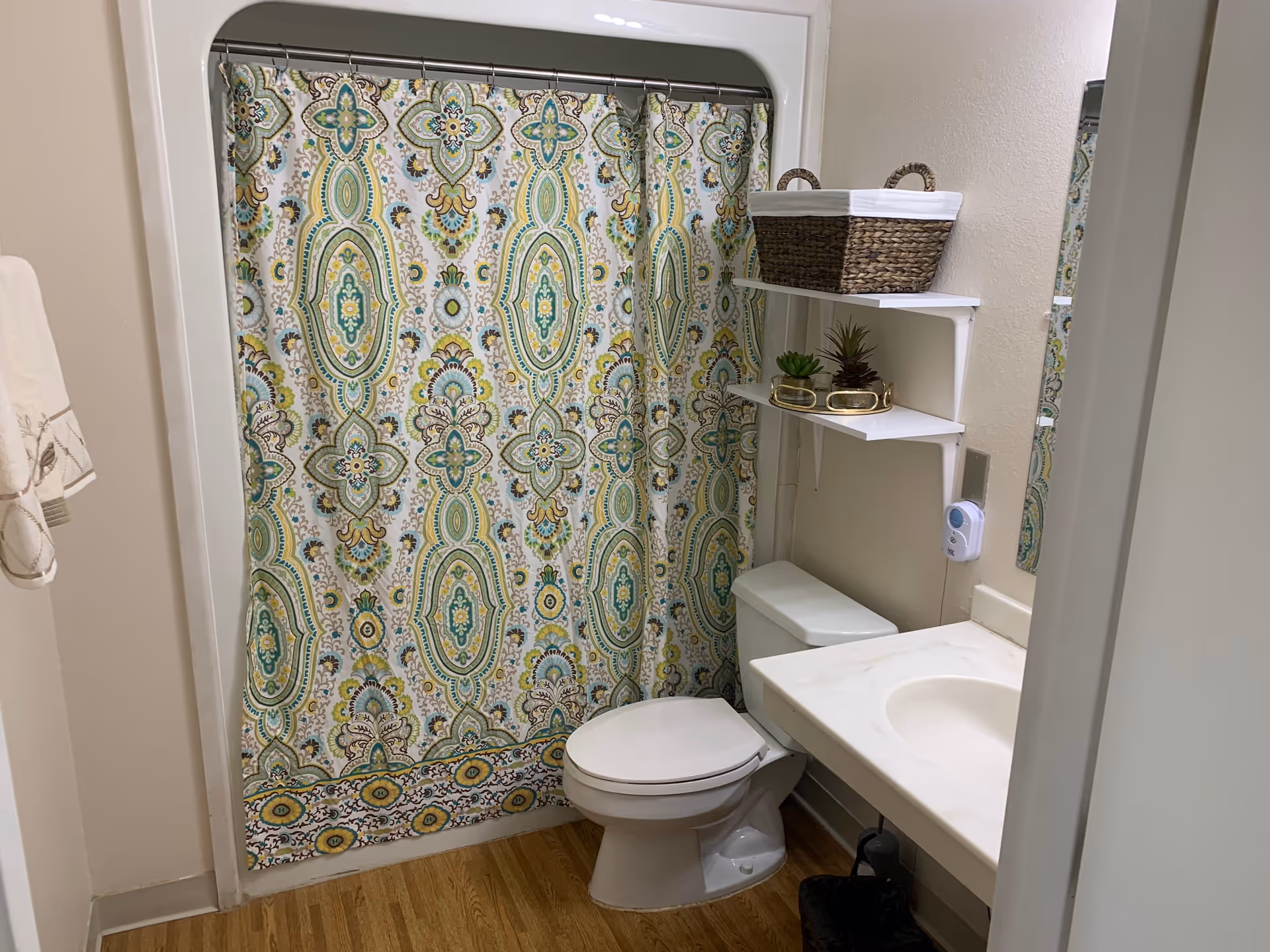 A small bathroom with a toilet, a sink with a countertop, and a shower with a decorative curtain featuring a green, blue, and yellow pattern. Above the toilet, there are two white shelves holding a wicker basket and small potted plants. A towel hangs on the left wall.