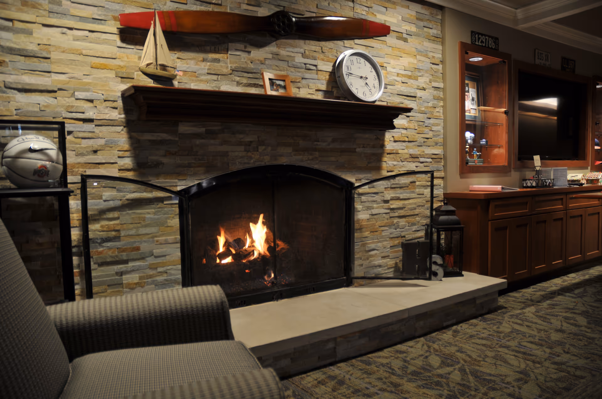 A cozy living room area with a stone fireplace featuring a burning fire. Above the fireplace mantel, there is a small model sailboat, a framed photo, a clock, and a wooden oar mounted on the stone wall. To the right, there is a wooden cabinet with a television and display shelves. In the foreground, part of a cushioned armchair is visible.