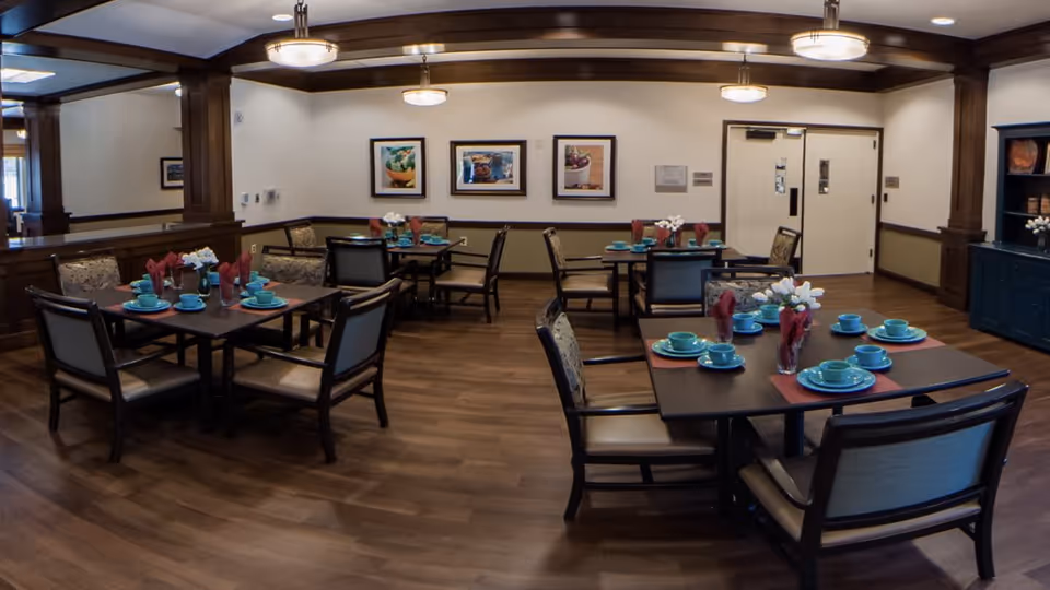 A dining room in a senior living facility with several square tables set with teal plates, cups, and red napkins. Each table has a small floral centerpiece. The room features wooden flooring, wood trim, framed artwork on the walls, and overhead lighting fixtures.