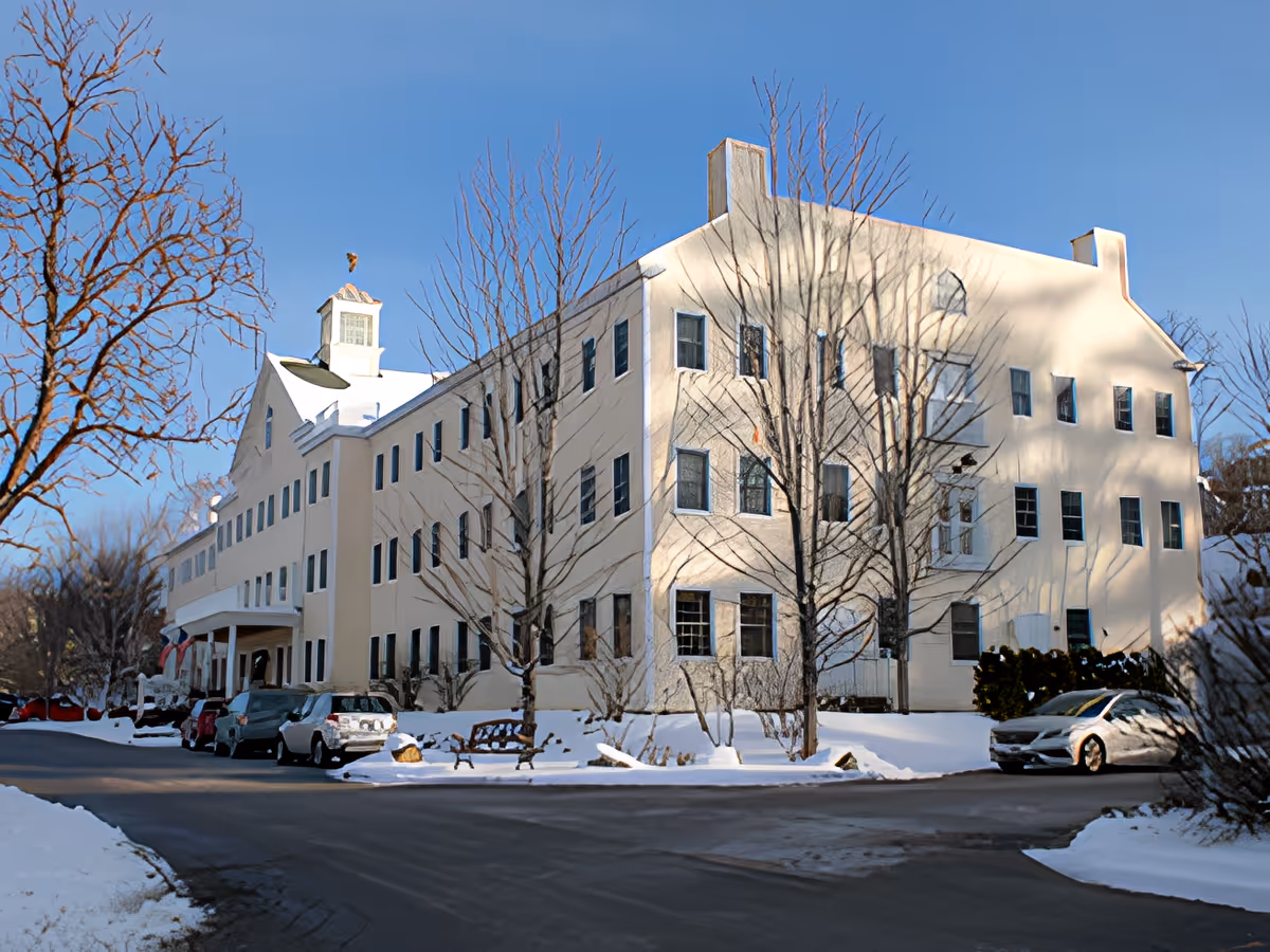 Exterior view of a large, multi-story beige building with many windows, surrounded by leafless trees and snow-covered ground. Several cars are parked along the driveway in front of the building under a clear blue sky.