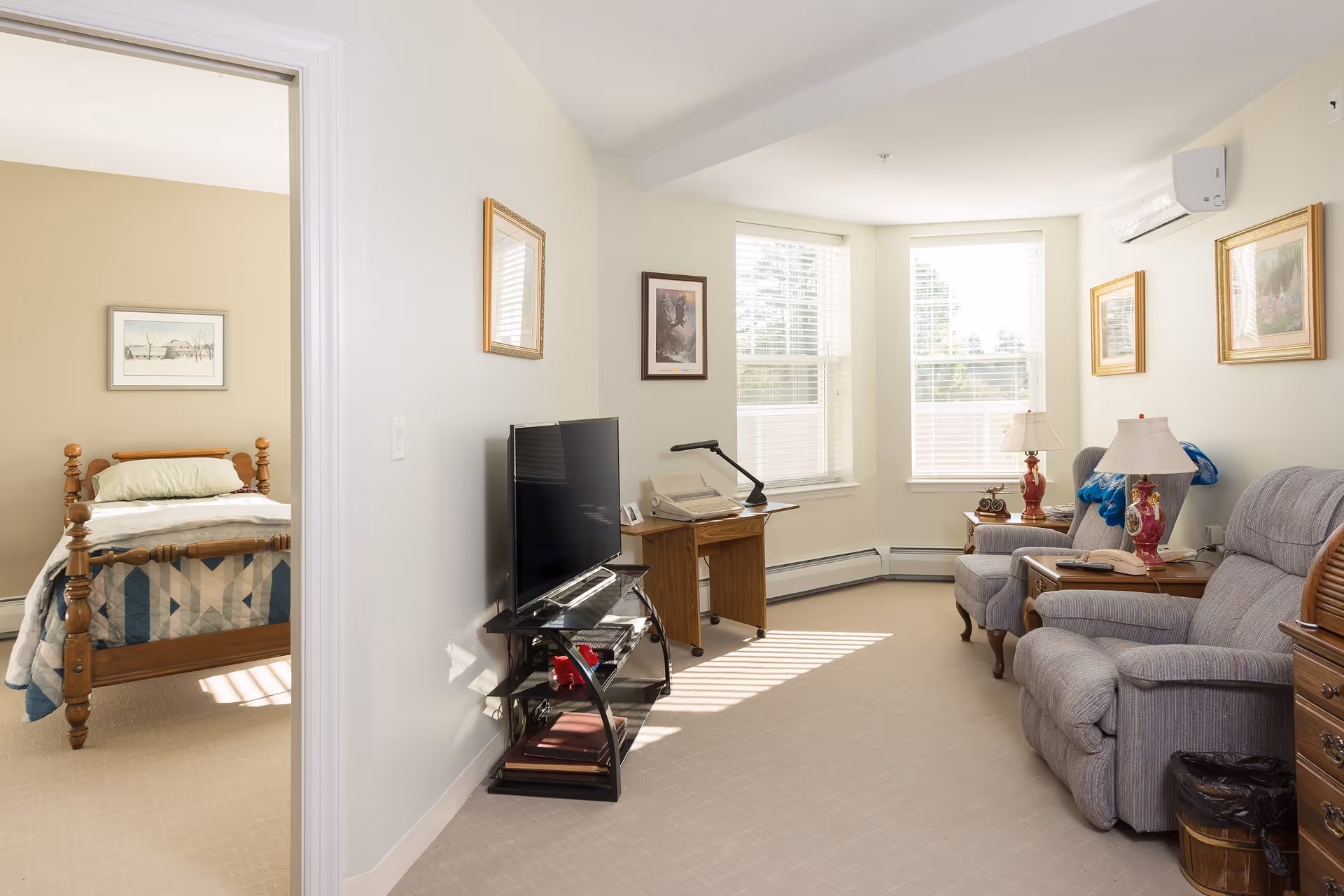 A bright living room with two upholstered armchairs, side tables with lamps, a TV on a stand, and a small wooden desk with a lamp and typewriter. The room has large windows with white blinds letting in natural light. A bedroom with a wooden bed and quilt is visible through an open doorway.