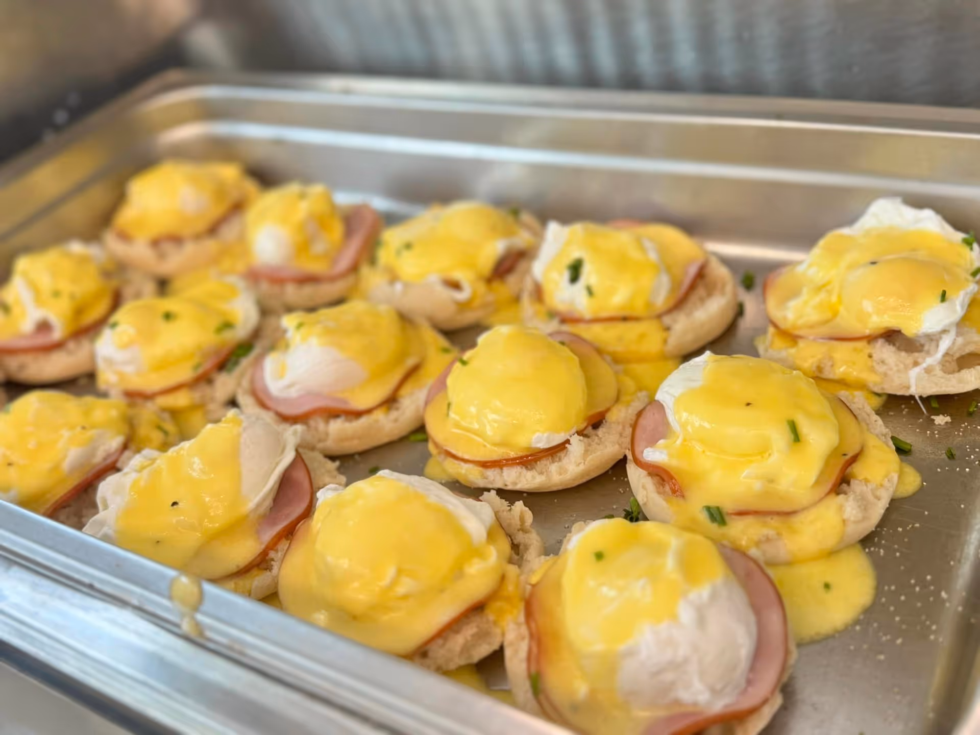 Tray filled with multiple servings of Eggs Benedict, each consisting of an English muffin topped with ham, a poached egg, and hollandaise sauce, garnished with chopped herbs.