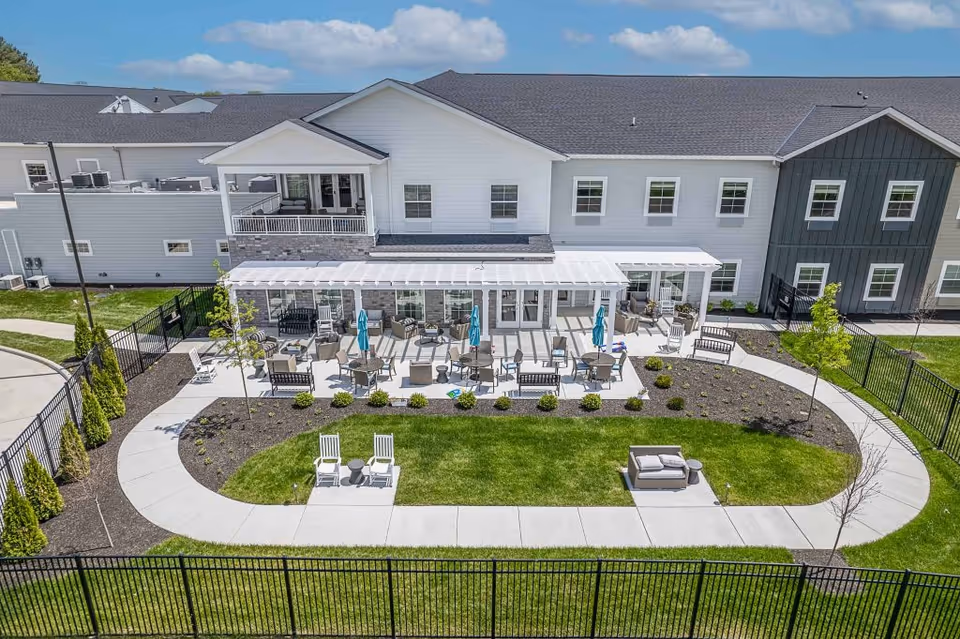 Outdoor patio area at Provision Living at Crown Ridge featuring multiple tables with chairs and umbrellas, benches, lounge chairs, and a well-maintained lawn surrounded by a curved sidewalk and black metal fencing. The building exterior is visible in the background under a partly cloudy sky.