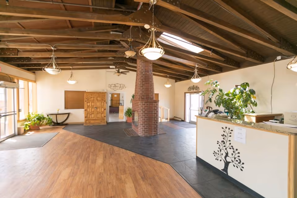 Spacious interior lobby area with wooden beams on the ceiling, a central brick fireplace, hanging light fixtures, a reception desk with a decorative tree design, potted plants, and large windows letting in natural light.
