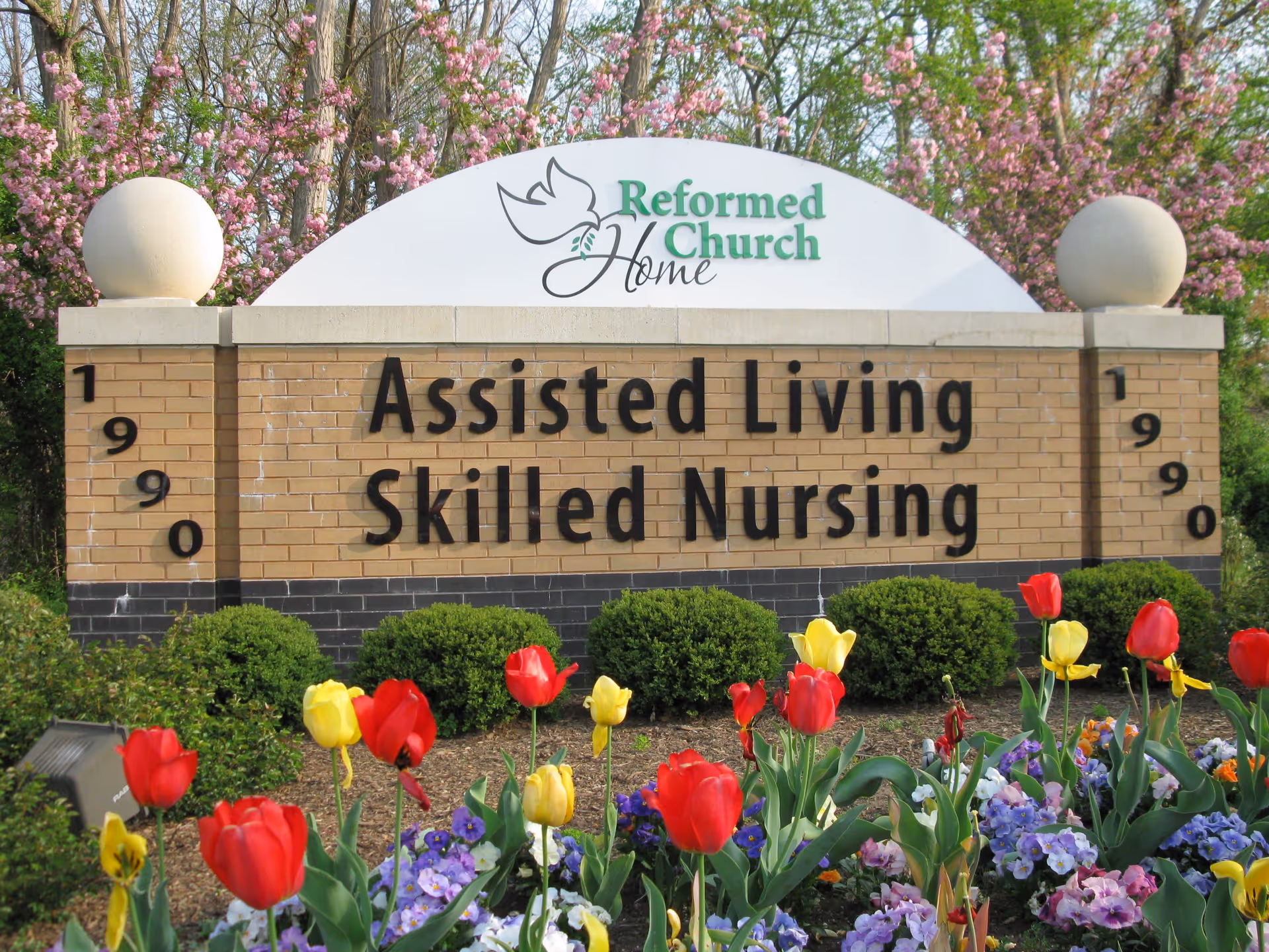 A brick sign for Reformed Church Home with the text 'Assisted Living Skilled Nursing' surrounded by blooming flowers including red and yellow tulips and purple pansies, with trees and pink blossoms in the background.