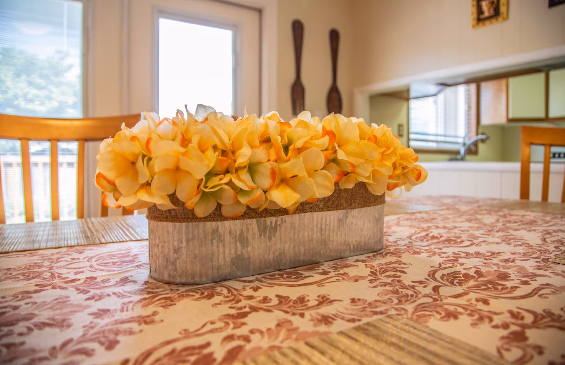 A dining table with a yellow floral centerpiece, wooden chairs, and a view into the adjacent kitchen.