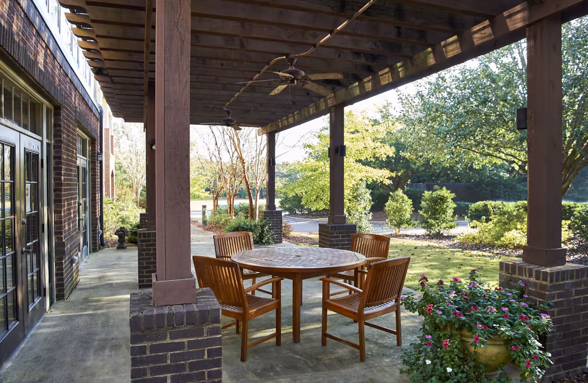 Outdoor covered patio area with a round wooden table and four wooden chairs. The patio has brick pillars supporting a wooden pergola roof with ceiling fans. Surrounding the patio are green bushes, trees, and a flower pot with pink and white flowers.