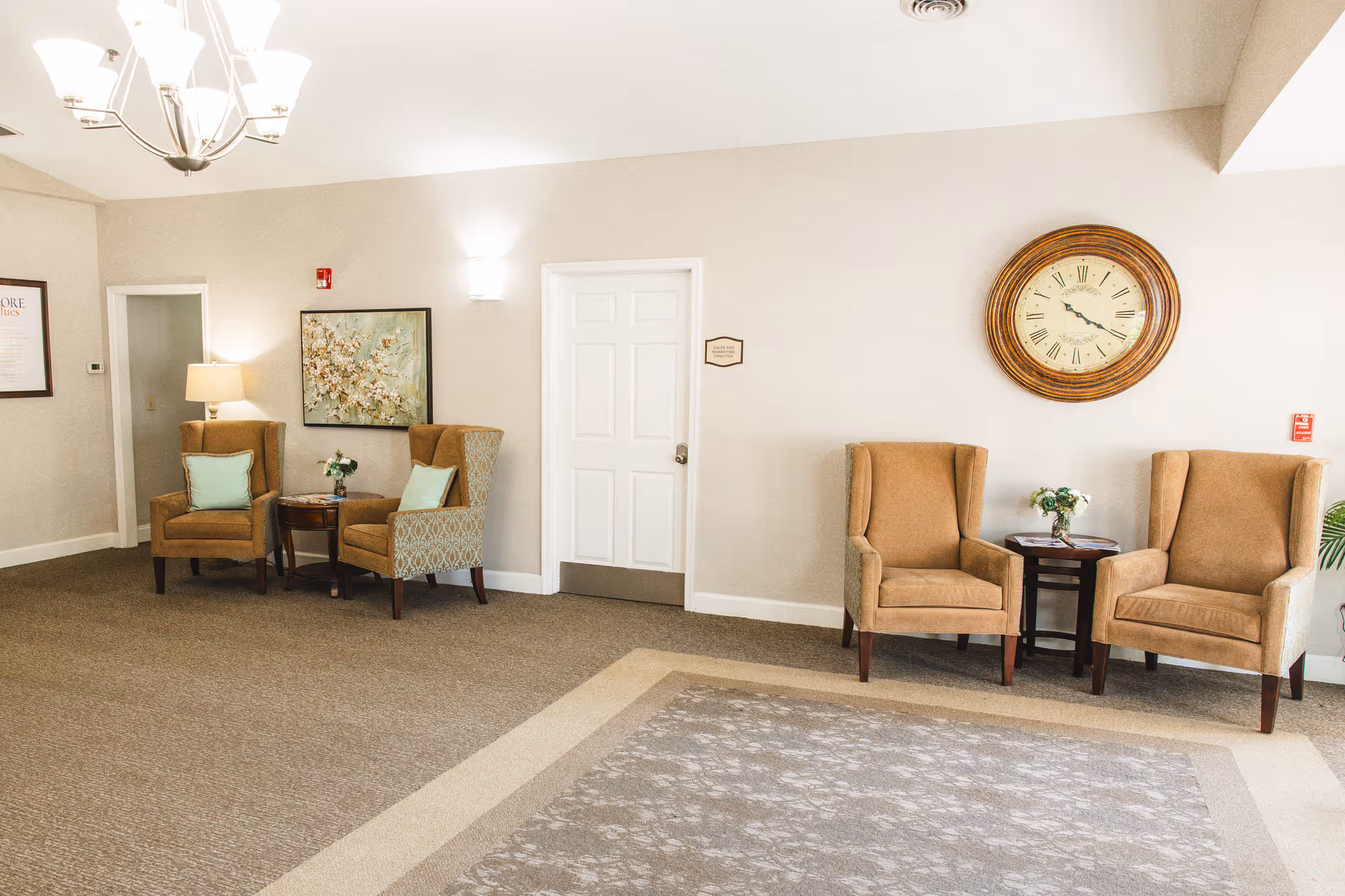 A well-lit sitting area in a senior living facility with four beige armchairs arranged in pairs around two small dark wooden tables. Each table has a small flower arrangement. The walls are light-colored, decorated with a large round clock and a floral painting. There is a white door in the center of the wall and a chandelier hanging from the ceiling.