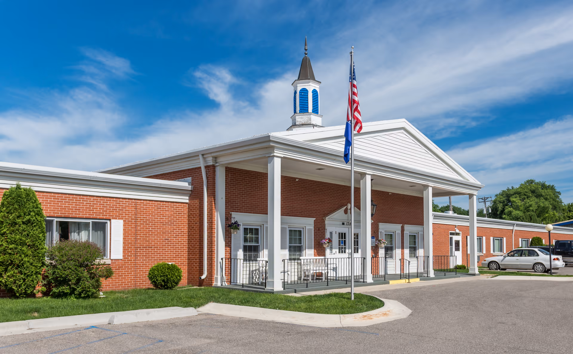 Front entrance of a brick senior living facility with white columns, flagpoles, a small cupola, and parked cars under a blue sky.
