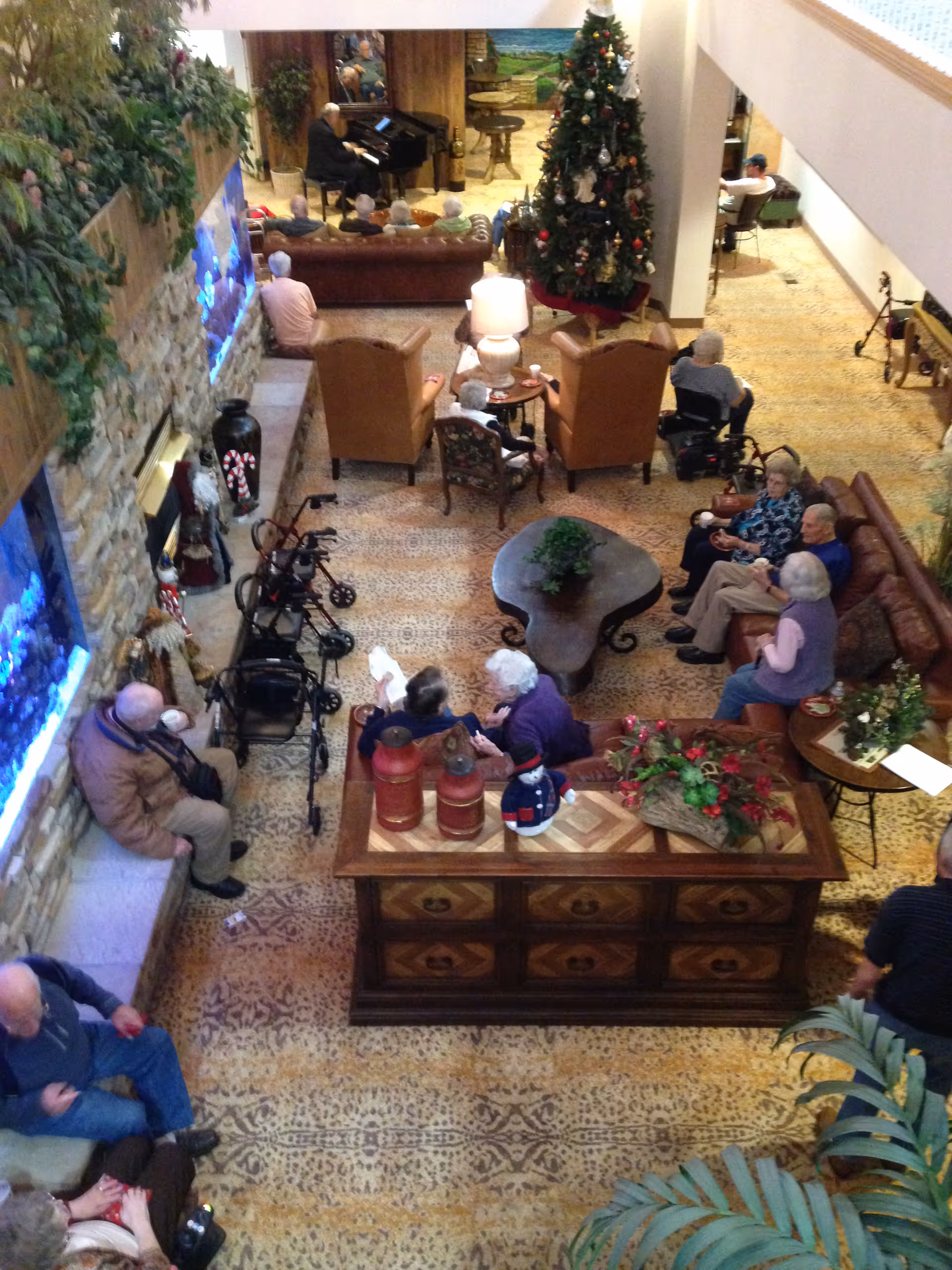 Overhead view of a senior living facility common area with elderly residents sitting on sofas and chairs, some using walkers. A decorated Christmas tree is visible near the center, and a person is playing a piano in the background. The room has a patterned carpet, stone wall with aquariums, and various plants and decorations.