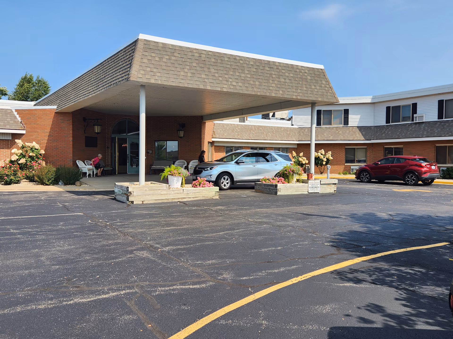 Entrance of Brandon Wood Retirement Center showing a covered drop-off area with two cars parked nearby. There are flower beds with blooming flowers and two people sitting on chairs near the entrance. The building is made of brick with some white siding and has multiple windows.
